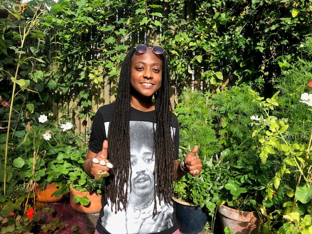 Black woman with long braids, sunglasses, funky rings and a big grin gives us a thumbs up. She's in a sunny garden.