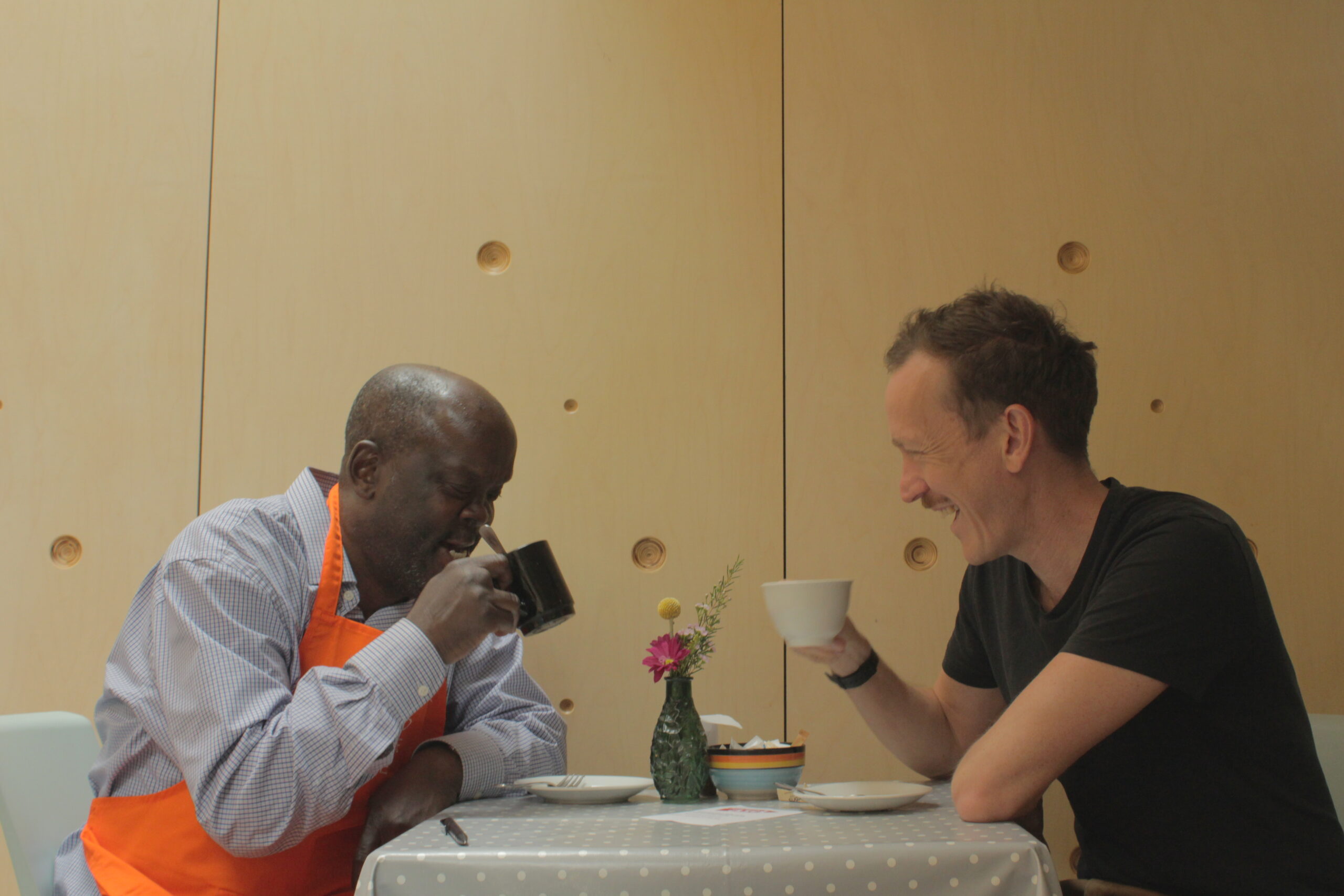a Black man in an orange apron sitting at a table with a cup of coffee and a white man with a moustache laughing