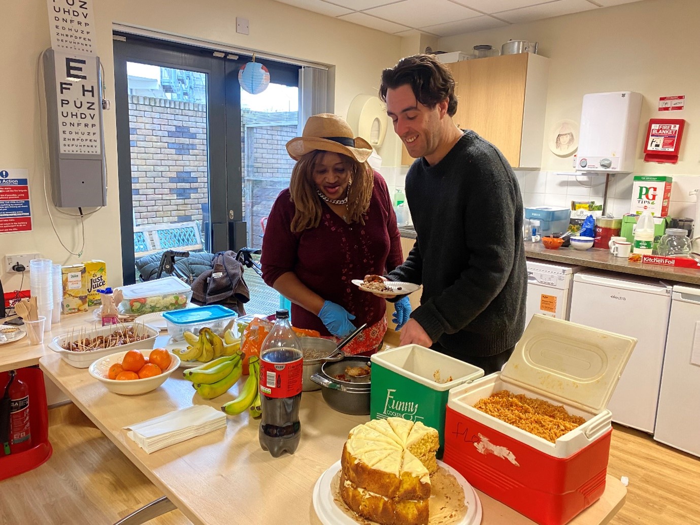 Black woman in a straw hat and kitchen gloves is smiling and chatting to a younger White man holding a plate of food. They're laughing and chatting over a table full of food.