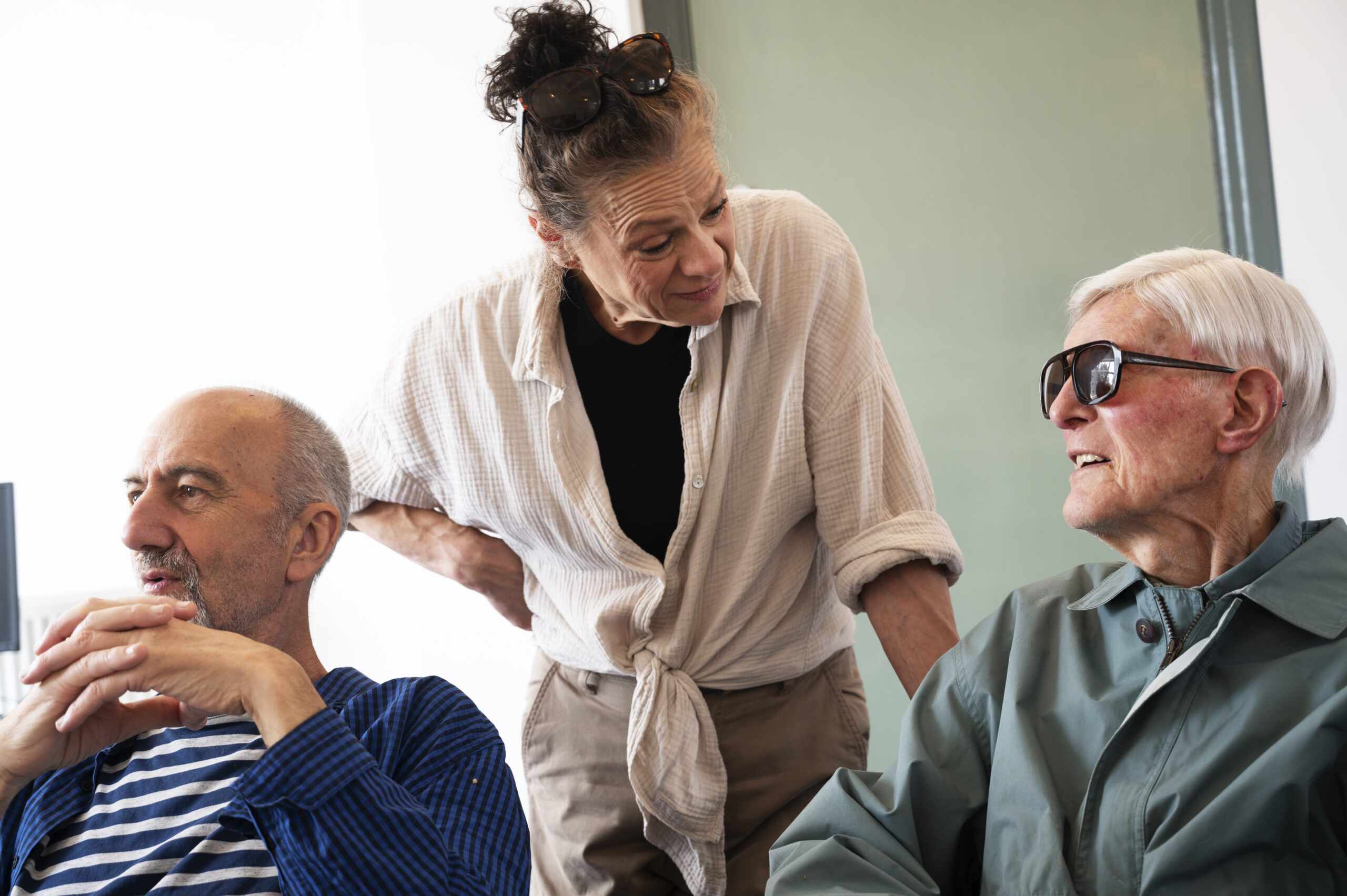 a woman with light skin smiling and chatting to a seated older man with light skin and sunglasses